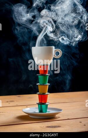 Tazza di caffè in cima a una torre fatta con cialde di caffè colorate e utilizzata su un tavolo di legno su sfondo nero. Foto Stock