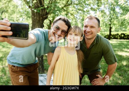 Coppia gay che fa ritratto selfie sul telefono cellulare insieme con la loro figlia adottiva all'aperto Foto Stock