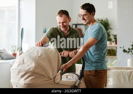 Felice giovane coppia gay in piedi in camera con passeggino che adottano il bambino Foto Stock
