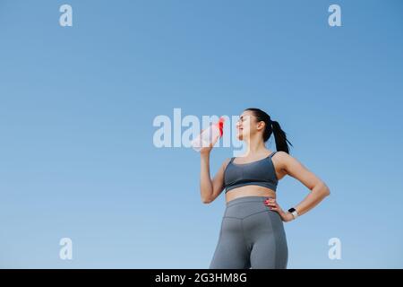 Giovane donna sorridente in abbigliamento sportivo che beve acqua. Sopra il cielo blu Foto Stock