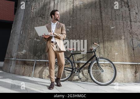 tutta la lunghezza di uomo d'affari allegro in auricolari che tengono il laptop vicino all'edificio e bicicletta all'esterno Foto Stock