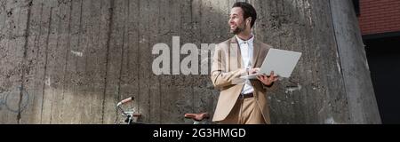 uomo d'affari allegro in auricolari che tengono il laptop vicino a edificio esterno, banner Foto Stock