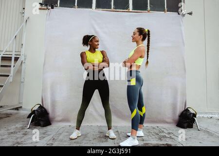 Due donne in abbigliamento sportivo che si guardano l'una all'altra sul tetto. Donne sportive con braccia incrociate in piedi insieme e sorridenti. Foto Stock