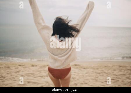 Immagine offuscata di spensierata bella donna in maglia maglione che corre su spiaggia sabbiosa a mare freddo, divertendosi. Concetto di vacanza estiva. Elegante giovane ha Foto Stock