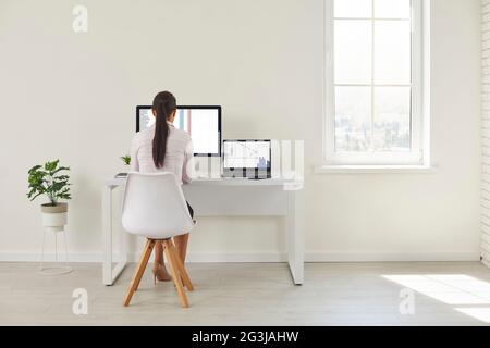 Vista sul retro di una donna seduta sulla scrivania dell'ufficio e che lavora su un computer desktop e un computer portatile Foto Stock