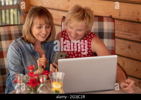 Due donne senior che usano il computer portatile a casa, sulla veranda di casa in legno Foto Stock