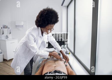 Concetto di medicina, salute e controllo del cuore. Giovane donna afro-americana medico con apparecchiatura di elettrocardiogramma che fa il test del cardiogramma al paziente maschio in ospedale, fissando gli elettrodi sul torace del paziente Foto Stock