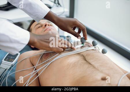 Immagine ritagliata di una donna afroamericana medico o infermiera attaccando holter e elettrodi a vuoto sul petto di un giovane paziente di sesso maschile in ospedale. Concetto di ECG e salute del cuore. Controllo annuale dello stato Foto Stock