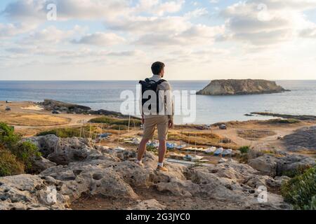 L'uomo con lo zaino sta camminando lungo una costa rocciosa al tramonto vicino al Mar Mediterraneo a Cipro. Il turista maschile ammira la vista Yeronisos, o Santo Foto Stock