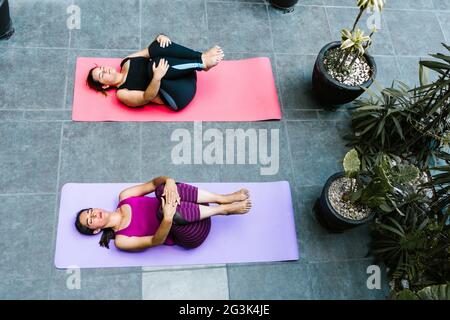 Donne latine che fanno yoga sul pavimento con le ginocchia al petto e gli occhi chiusi sulla terrazza a casa Foto Stock