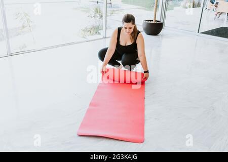 Ragazza latina che srotola il tappetino yoga sul pavimento vicino alla finestra di casa Foto Stock