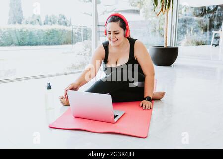 Ragazza ispanica curvy con cuffie rosse e abbigliamento sportivo che porta una lezione di yoga online su un computer portatile a casa Foto Stock