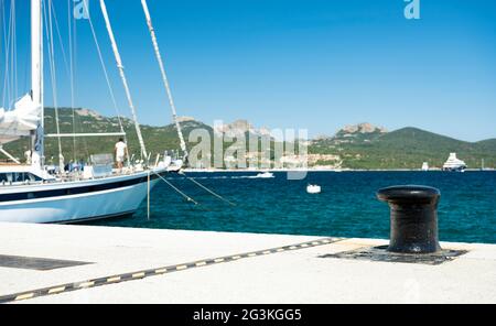 (Fuoco selettivo) barca a vela sfocata ormeggiata a un porto durante una giornata di sole con un bollard in primo piano. Porto Rotondo, Sardegna, Italia. Foto Stock
