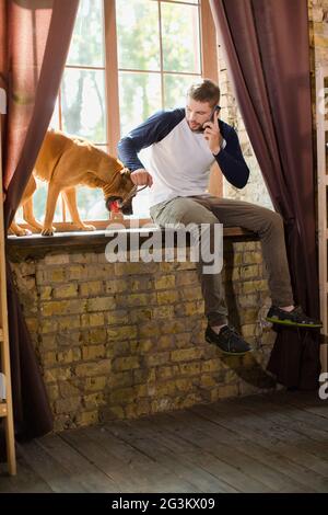 View of young man playing with dog and talking on phone. Foto Stock
