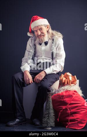 Vecchio Santa stanco seduto su sedia con borsa regalo nelle vicinanze. Foto Stock