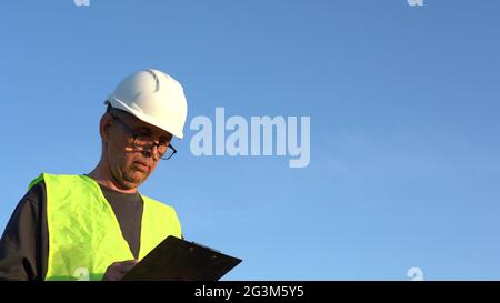 Un uomo anziano che indossa occhiali e un casco da lavoro fa note contro un cielo blu chiaro Foto Stock