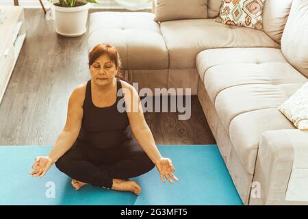 Donna di mezza età in abbigliamento sportivo in posizione yoga - donna anziana si rilassa meditando in casa sua - concetto di vita sana e benessere - filtro caldo o Foto Stock