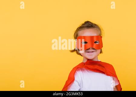 Bambina bionda vestita come supereroina supereroe con capo e maschera rossa, sorridente, su sfondo giallo Foto Stock