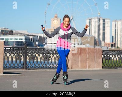 La giovane donna sta rollerblading con uno smartphone nelle sue mani e ascoltando la musica attraverso le cuffie. Si sposta alla musica e gode la vita. Foto Stock