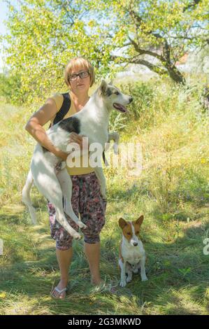 Bel ritratto esterno di donna Ucraina matura prendendo sulle mani grande cane di razza bianca mista, mentre basenji uno seduto accanto in frutteto albicocca a su Foto Stock