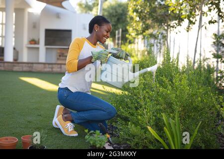 Sorridente afroamericano donna giardinaggio, inginocchiandosi in giardino innaffiando piante con annaffiatoio lattina Foto Stock