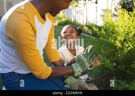 Sorridente madre afroamericana e figlia inginocchiati tendendo a piante in giardino soleggiato Foto Stock