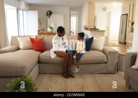 Sorridente donna afroamericana medico in visita ragazza paziente a casa, seduta sul divano che parla Foto Stock