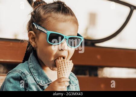 Una ragazza in un vestito di denim e gli occhiali da sole mangia il gelato su una panchina del parco. Foto di alta qualità Foto Stock