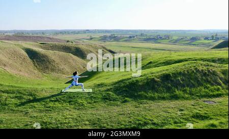 Yoga femminile praticando yoga in natura, verdi colline in estate su un tappetino yoga. Una ragazza caucasica dai capelli castani si trova in un'asana con abbigliamento sportivo, leggings e una T-shirt con capelli intrecciati in una coda di pony. Foto Stock