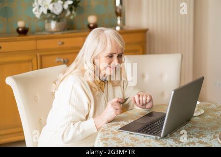 Sorridente donna anziana usa il laptop e la carta di credito mentre si siede al tavolo Foto Stock