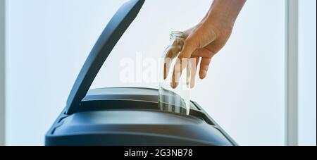 closeup di un giovane uomo caucasico che lancia una bottiglia di vetro vuota nel cestino di riciclaggio della casa, in un formato panoramico da utilizzare come banner web o intestazione Foto Stock