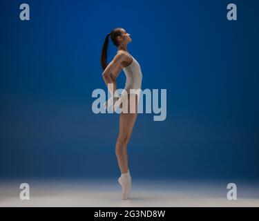 Sulle punte delle dita dei piedi. Giovane ballerina bella in azione isolata su sfondo blu. Foto Stock