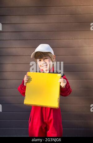 ragazzo di 4- 5 anni in uniforme meccanica rossa e tappo sullo sfondo di una porta del garage. Capretto tiene la scatola gialla davanti a lui, su cui ci è posto f Foto Stock