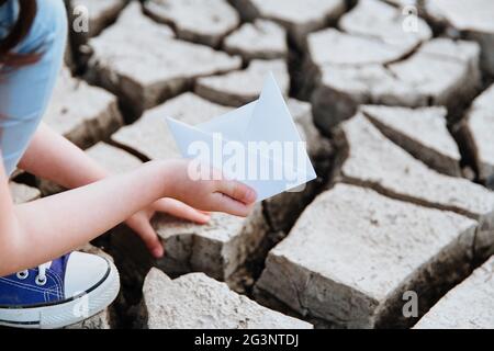 La ragazza abbassa la barca di carta sul terreno asciutto e incrinato. Crisi idrica e concetto di cambiamento climatico. Riscaldamento globale Foto Stock