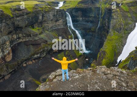Vista aerea dell'uomo con giacca gialla alza le mani godendoti la valle dell'altopiano islandese e la cascata. Islanda Foto Stock