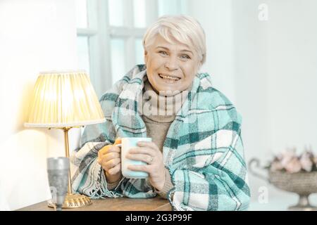 Sorridente Old Lady avvolto in un Checkered Plaid sta tenendo UNA tazza di tè. Foto Stock