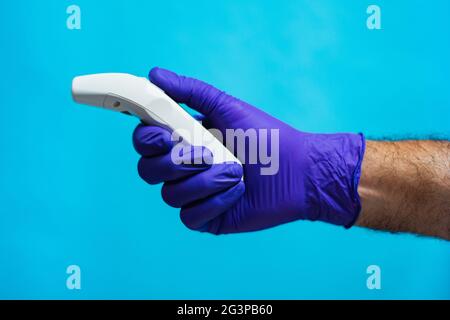 Una mano del medico che indossa un guanto viola che tiene un termometro su sfondo blu. Protezione Corona-virus. (Vista laterale) Foto Stock