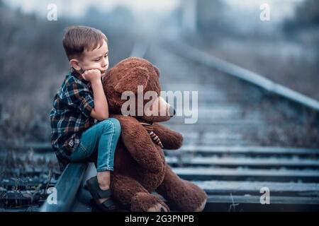 Un ragazzino con un grande orsacchiotto si siede sulle rotaie della ferrovia. Foto Stock