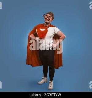 L'uomo super eroe romantico tiene il grande cuore rosso. Uomo felice innamorato con costume da supereroe e occhiali a forma di cuore isolati su bl Foto Stock