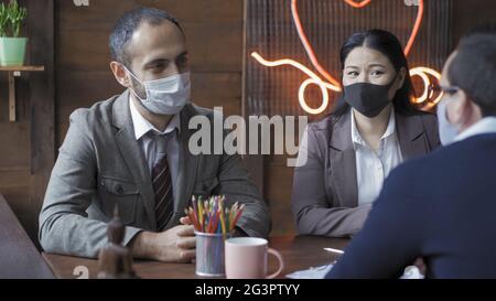 Riunione del team aziendale in ufficio. I colleghi sono cordiali e comunicano mentre si siedono alla scrivania dell'ufficio, indossando indumenti da lavoro e indumenti protettivi Foto Stock