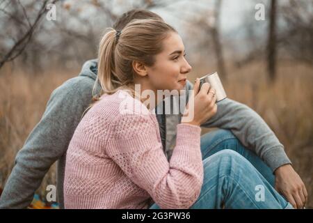 Picnic in natura. La giovane donna e l'uomo si riposano bevendo caffè caldo o tè mentre si siedono sulla coperta all'erba d'autunno all'aperto. Profil Foto Stock