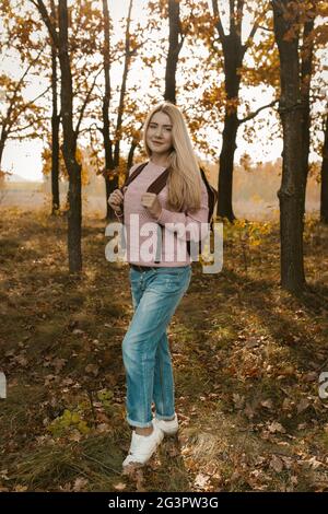 Donna viaggiatore in posa con uno zaino sullo sfondo della foresta autunnale retroilluminata dai raggi del sole. Bellissimo viaggio biondo Foto Stock