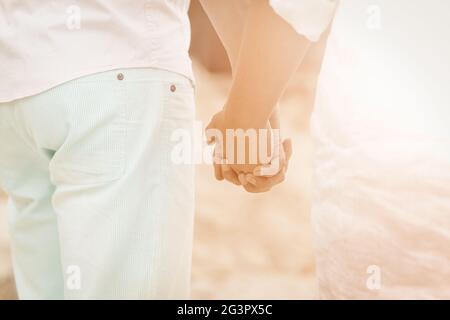 Coppia che tiene le mani al mare. Vista posteriore uomo e donna in abiti luminosi che tengono le mani camminano insieme lungo la spiaggia su un sole Foto Stock