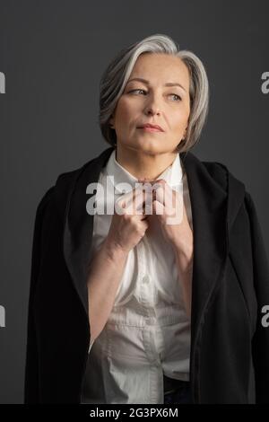 Affascinante donna dai capelli grigi guarda con attenzione il colletto di regolazione della camicia bianca, gettando una giacca nera sulle spalle. È Foto Stock