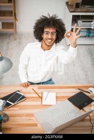 L'arabo mostra un bel gesto che sorride alla fotocamera. Uomo felice in occhiali che lavora a casa al ritmo di lavoro con computer portatile, telefono cellulare, non Foto Stock