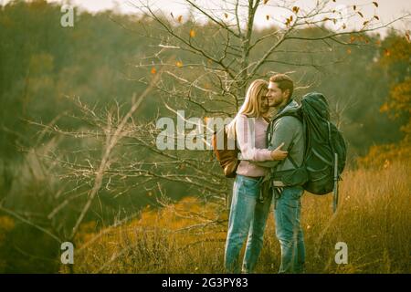 Felici sposi di natura in piedi con zaini su erba d'autunno. Giovane uomo caucasico e donna sorridono abbracciando mentre enjo Foto Stock