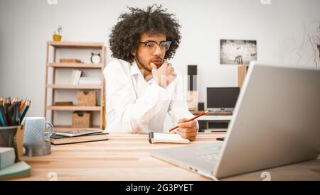 Il giovane pensivo è impegnato a casa davanti al computer. Studente arabo o uomo d'affari che prende appunti in un notebook. Educatio domestico Foto Stock