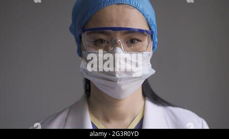 Medico in occhiali di sicurezza e maschera. Ritratto di donna asiatica in uniforme medica e occhiali di protezione guardando la macchina fotografica su sfondo grigio Foto Stock