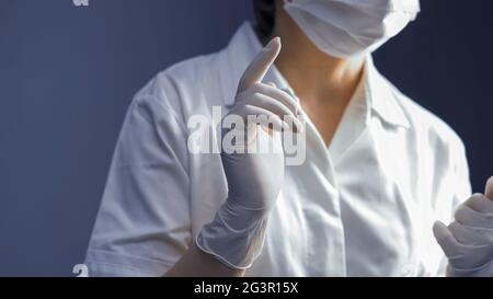 Donna che indossa guanti di gomma bianca e uniforme medica. Mettere a fuoco la mano femminile in primo piano. Primo piano. Concetto di igiene. TINTE Foto Stock