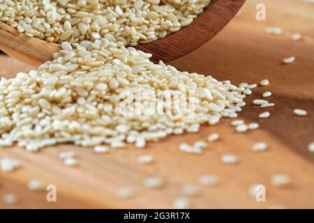 Semi di sesamo bianchi che versano da una piccola tazza a un tavolo di legno, dettaglio closeup Foto Stock